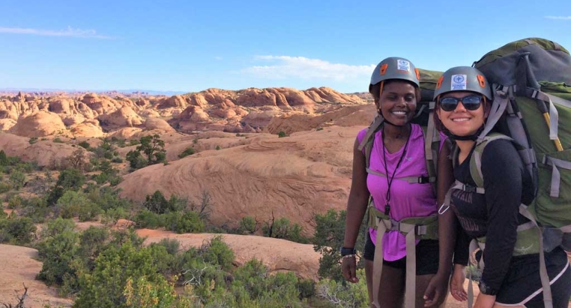 Two people wearing safety gear and carrying backpacks smile for a photo in a high desert landscape. 